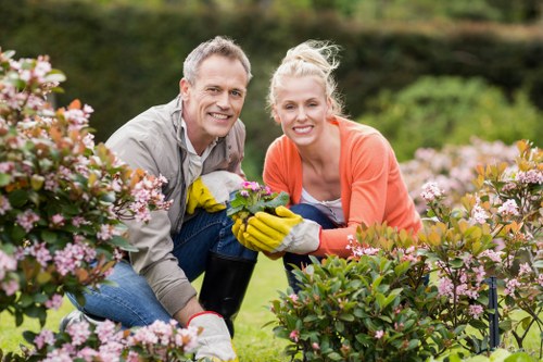 A man and a woman are kneeling together in a well-maintained garden, surrounded by blooming pink and purple flowering bushes and lush green foliage. The man, with short grey hair and wearing a grey shirt, is holding a gardening tool while smiling at the camera. The woman, with blonde hair tied back and dressed in an orange cardigan over a light top, is wearing bright yellow gardening gloves and is also smiling. In the background, there are small trees and a neatly trimmed lawn under a partly cloudy sky, suggesting a mild, clear day suitable for outdoor gardening activities. The garden features a combination of flower beds, evergreen shrubs, and a well-kept grassy area, indicative of a typical private outdoor space in Catford or nearby South London. This scene exemplifies gardening and outdoor maintenance activities promoted by Gardening Services Catford and underscores the importance of regular garden care and landscaping in residential settings.